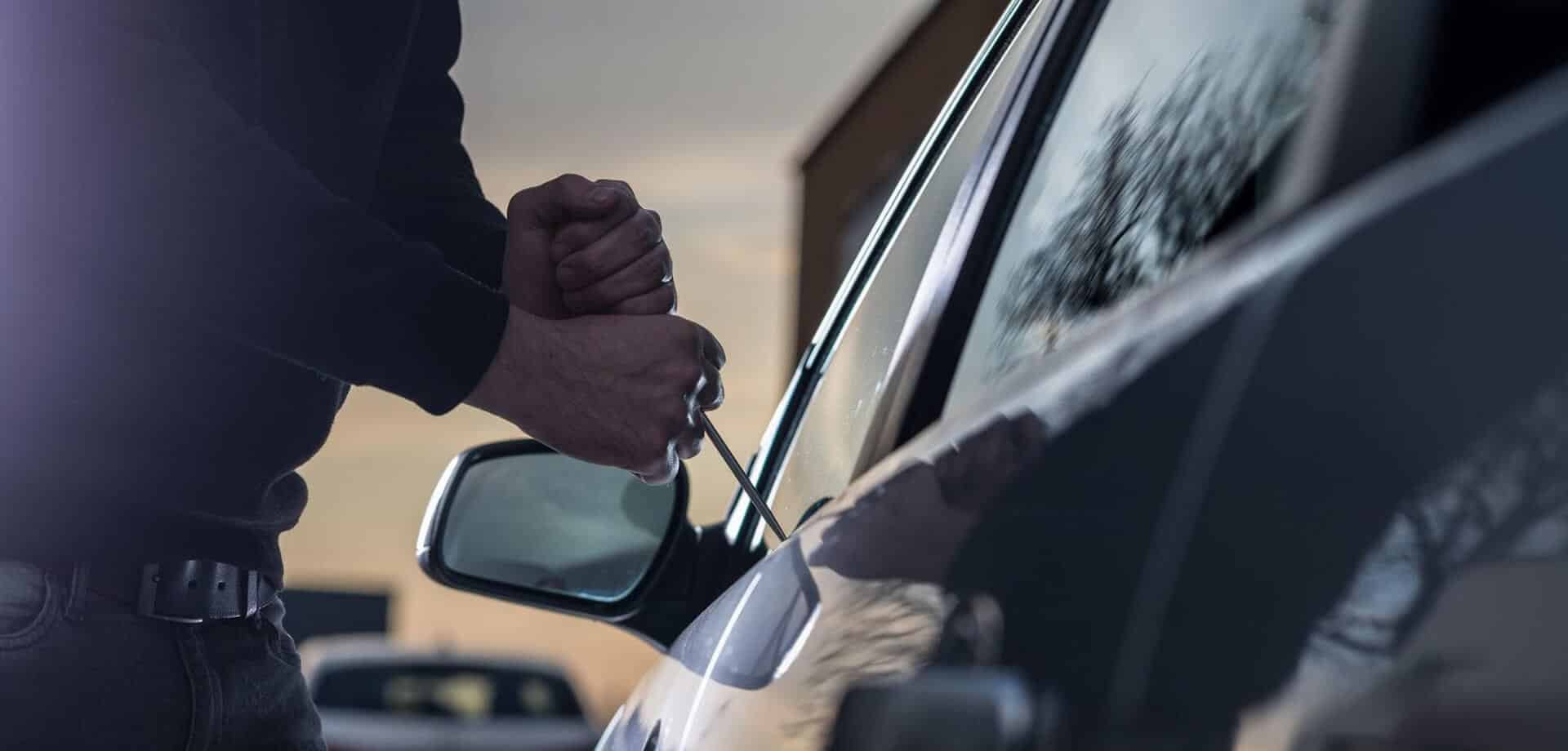 Person attempting to break into a car with a tool, representing vehicle theft prevention and remote immobilization technology using GPS and IoT solutions.