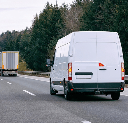White delivery van driving on a highway with a truck ahead, representing vehicle GPS tracking and fleet management solutions for logistics operations.
